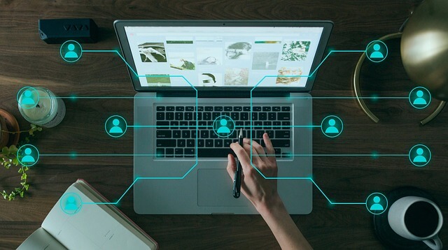 A imagem mostra um computador de cima com uma mão perto do teclado segurando uma caneta e diversos ícones de pessoas conectadas por linhas estão distribuídas por toda a imagem, representando as tendências de Marketing Digital 2023.